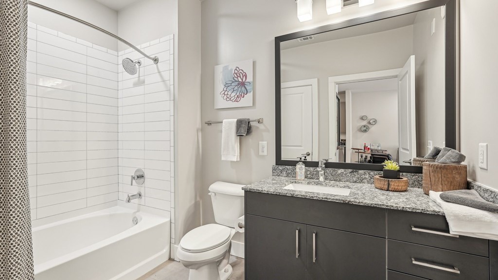 A bathroom with a white tub and shower, toilet, and granite countertop with a sink.