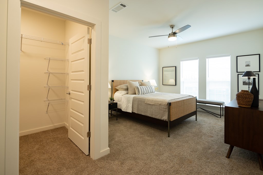 A bedroom with a bed, walk-in closet, dresser, windows, and a ceiling fan.