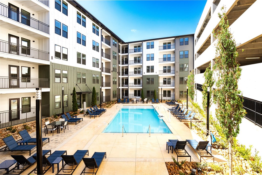 A swimming pool surrounded by lounge chairs and trees in front of apartment buildings.