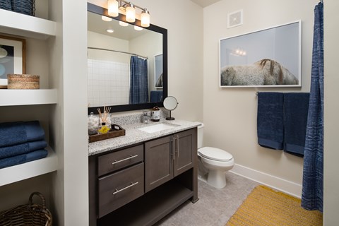 A bathroom with a toilet, sink, shower, shelving storage, and a mirror.