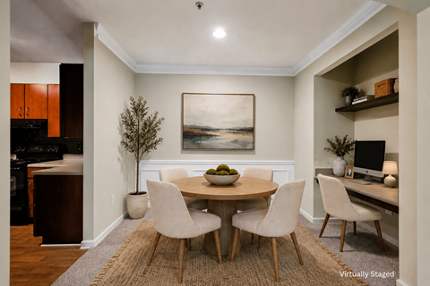 A dining room with a round table and chairs, a painting on the wall, and a kitchen area in the background.