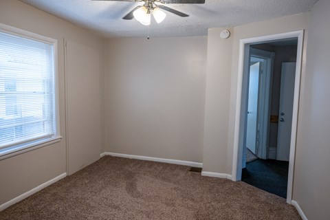 A room with a carpeted floor, a ceiling fan, and a door leading to another room.