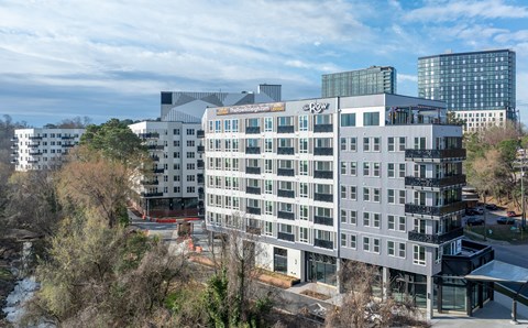 A large, modern building with many windows is surrounded by trees and other buildings.