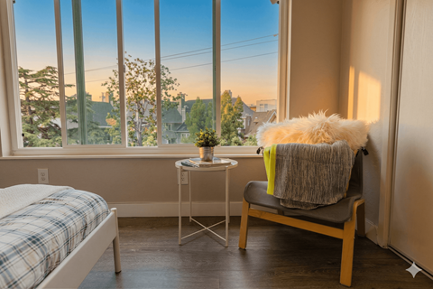A cozy bedroom with a bed, chair, and a view of the neighborhood through the window.