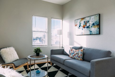 A living room with a grey couch, a black and white checkered floor, a small table with a plant on it, and a painting on the wall.