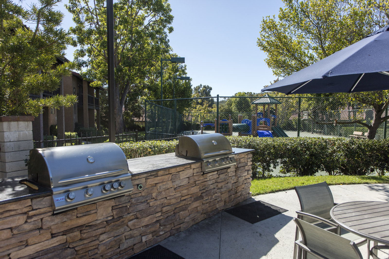 Apartments for Rent in Laguna Niguel CA - Pointe Niguel - BBQ Area with Two Grill Stations, Table, Chairs, Umbrella, Surrounding Trees, and View of Playground