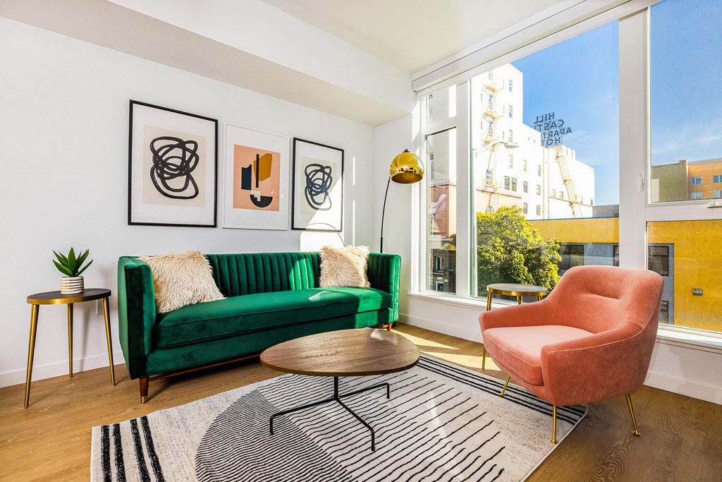 2 Bedroom Luxury Apartments in Downtown Oakland CA - Alice House - Living Room with Hardwood Style Flooring, a Green Sofa, Salmon Armchair, Wooden Coffee Table, Striped Rug, Golden Side Table and Standing Lamp, White Walls, and a Large Window with Urban Views
