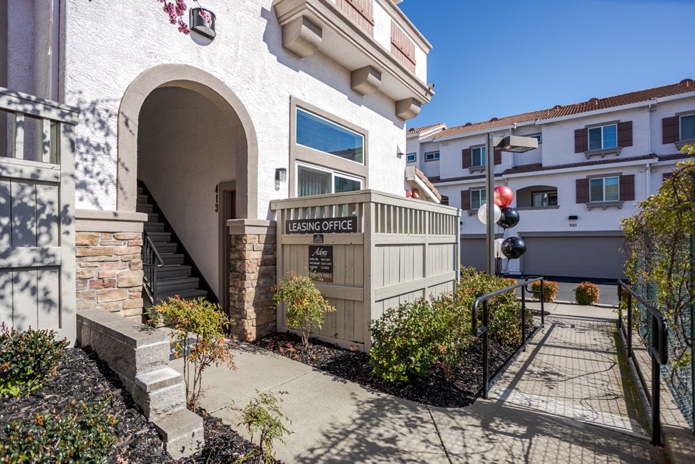 A building with a sign that says "LEASING OFFICE" is surrounded by plants and a sidewalk.