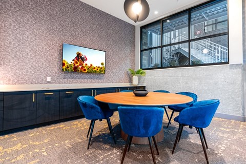 A room with a round table and blue chairs with a television on the wall.