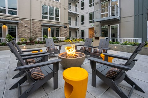 A fire pit surrounded by chairs and a yellow stool in front of a building.