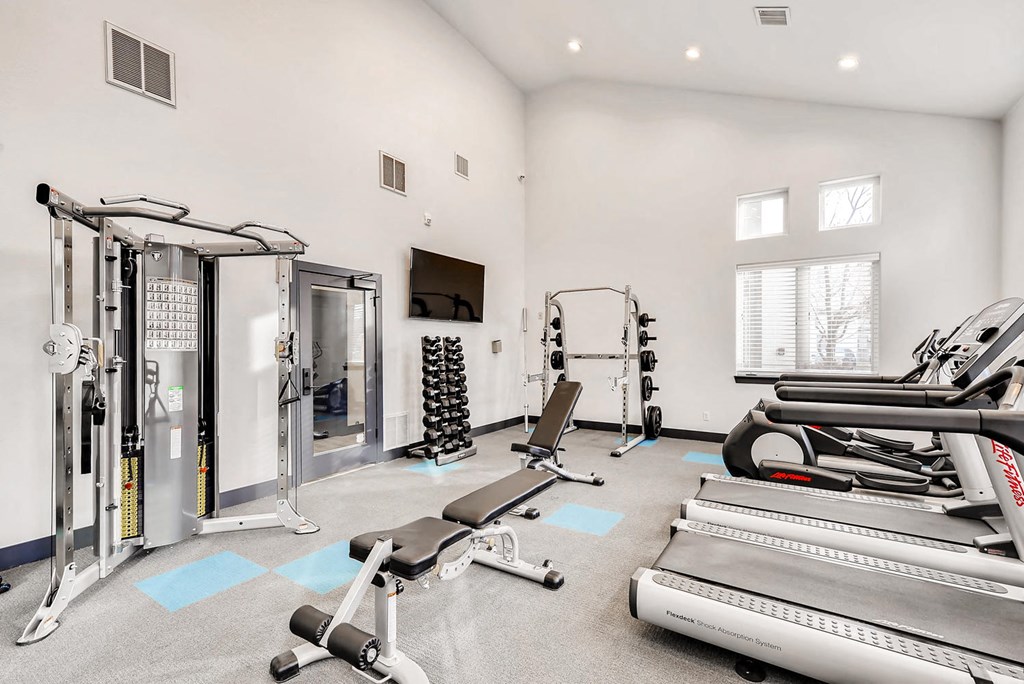 " Apartments Near Winsor, CO - Interior View Of Community Gym Which Features Two Running Treadmills, One Elliptical Machine, A Barbell Rack Under A TV, Smith Squatting Rack, And Cross-Training Machine