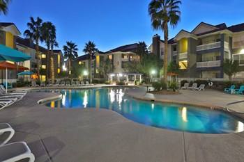 Apartments Phoenix AZ - Arboretum at South Mountain Apartments - Night Pool with Lounge Seating Surrounded by Palm Trees and Buildings - Photo Gallery 14