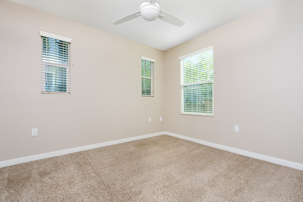 a bedroom with a ceiling fan and three windows, with carpeted floors.