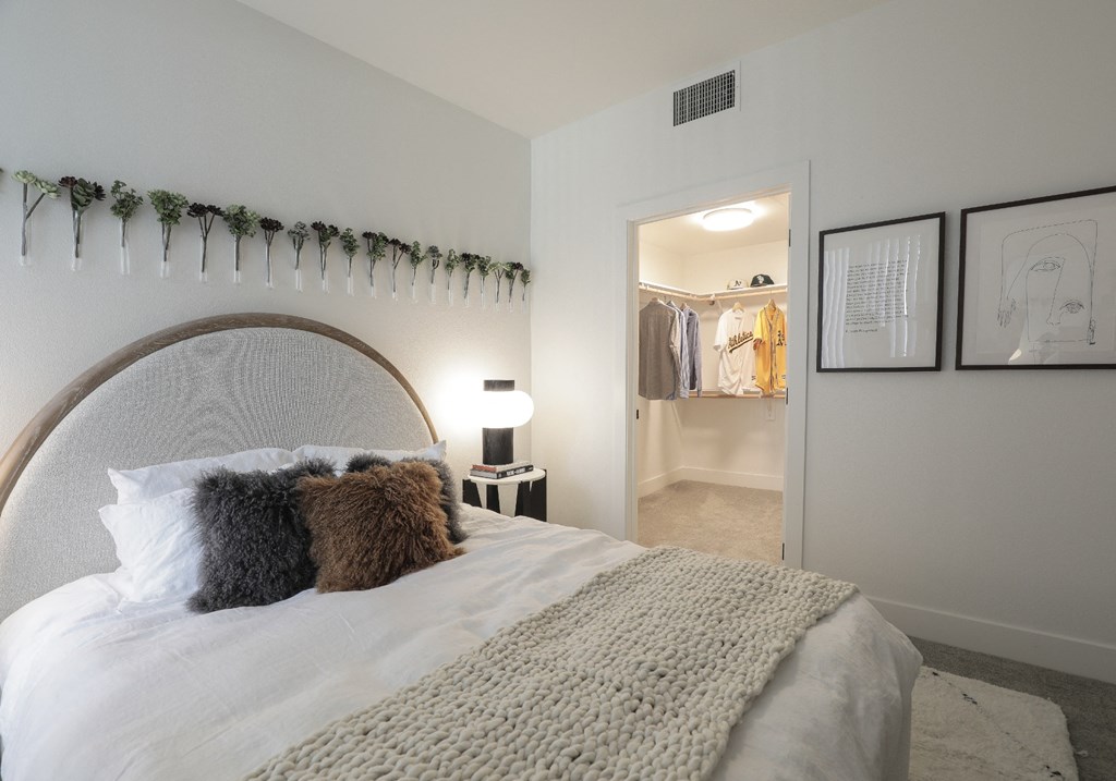 Apartments in Brooklyn Basin Oakland, CA - Artizan - Bedroom with White Walls, Plush Carpet Flooring, Large Bed, a Nighstand with Lamp, and Spacious Closet