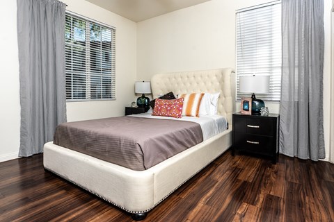 Apartments in Rancho Cucamonga, CA - Barrington Place - a Bedroom with Hardwood-Style Flooring, a Bed, Two Bedside Tables with Lamps, and Two Large Windows with Blinds and Curtains