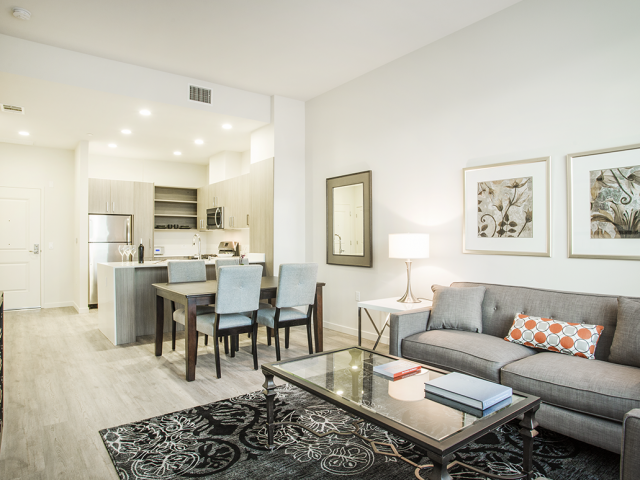 A modern living room with a grey couch, a glass coffee table, and a dining table with chairs.