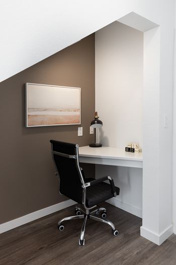 A black office chair is in front of a desk with a bottle of wine and a picture frame on it.