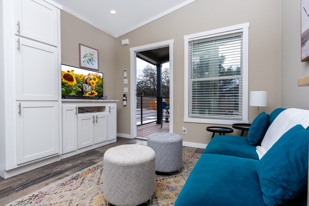 New Apartments In South Austin - Casata Austin - Spacious Living Room With Wood-Inspired Plank Flooring, Area Rug, Couch, Round Stools, Side Table With Lamp, High Ceilings, Built-In White Cabinets, TV, Window With Blinds, And Access To Patio