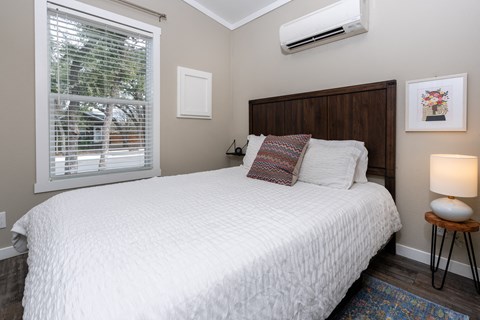 1-Bedroom Apartments in South Austin, TX - Casata Austin - Bedroom with Wood-Style Flooring, Area Rug, Bed with Wooden Headboard, Nightstand with Lamp, AC Unit, and Window with Blinds.