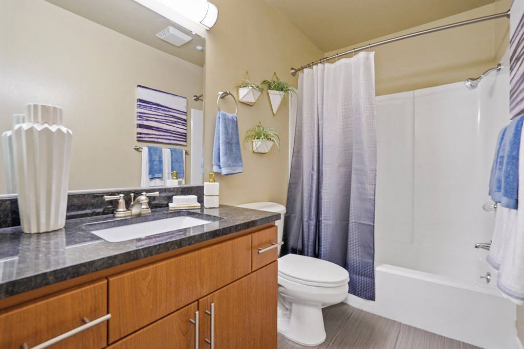 Apartments In Mountain View, CA - Americana Apartments - Bathroom With Large Vanity Mirror, Granite Countertop, Sink, Wood-Like Cabinets, Toilet, And A Shower With Tub at Americana Apartments, Mountai