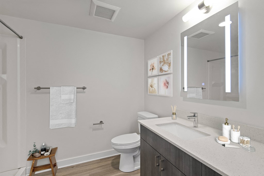 Downtown Seattle Apartments - Emmons On 3rd - Bathroom With Hardwood Flooring, Large Vanity Mirror With Lighting, Sink, Custom Cabinets, Toilet, And A Shower.