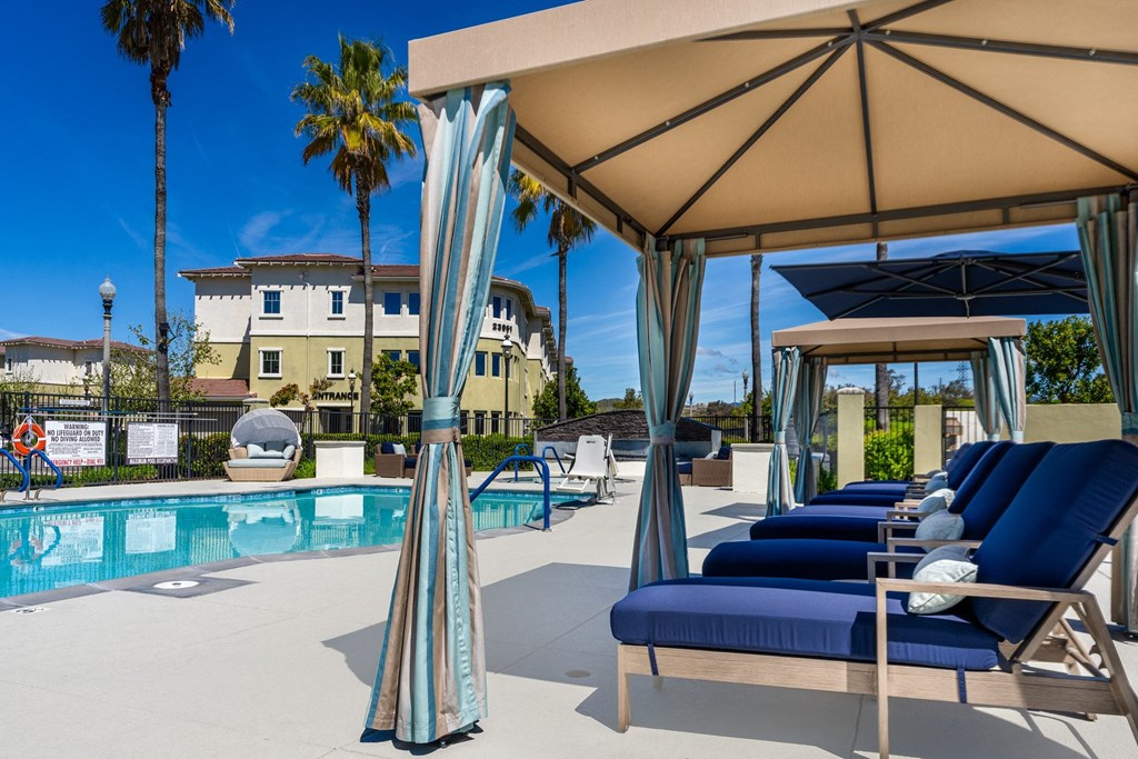 a pool with blue lounge chairs and a tan umbrella next to a pool