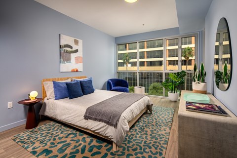 A bedroom with a bed, a blue chair, a rug, a potted plant, and a mirror.