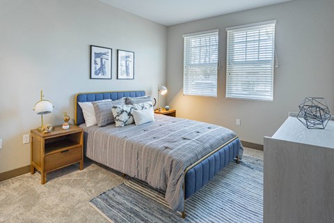 Studio Apartments In Burlingame, CA - Bayswater Burlingame - Spacious Bedroom With Plush Carpet Flooring, Area Rug, Bed, Nightstands With Lamps, Dresser, Wall Art, And Two Windows With Blinds at Baysw