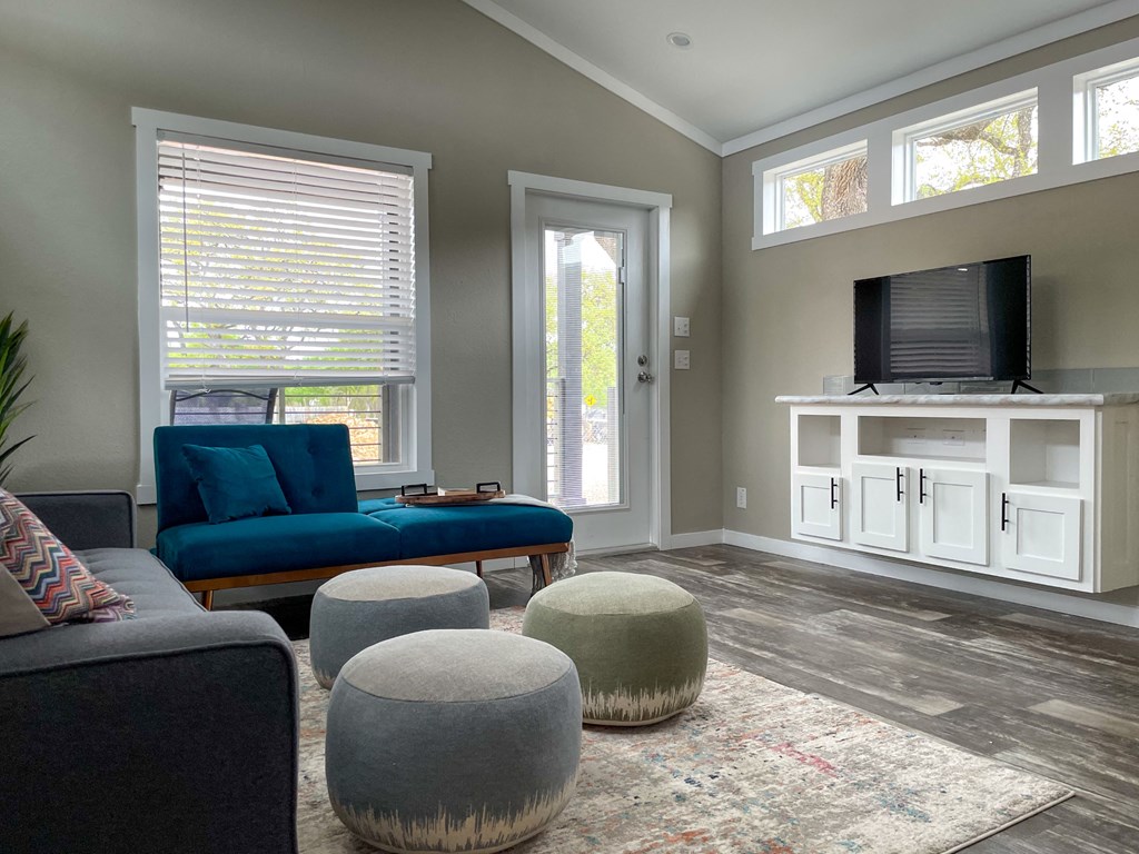 Brand New Apartments In South Austin, TX - Casata Austin - Living Room With Couch, Chair, Wood-Style Flooring, Three Ottomans, Area Rug, TV Stand With TV, And Five Windows.