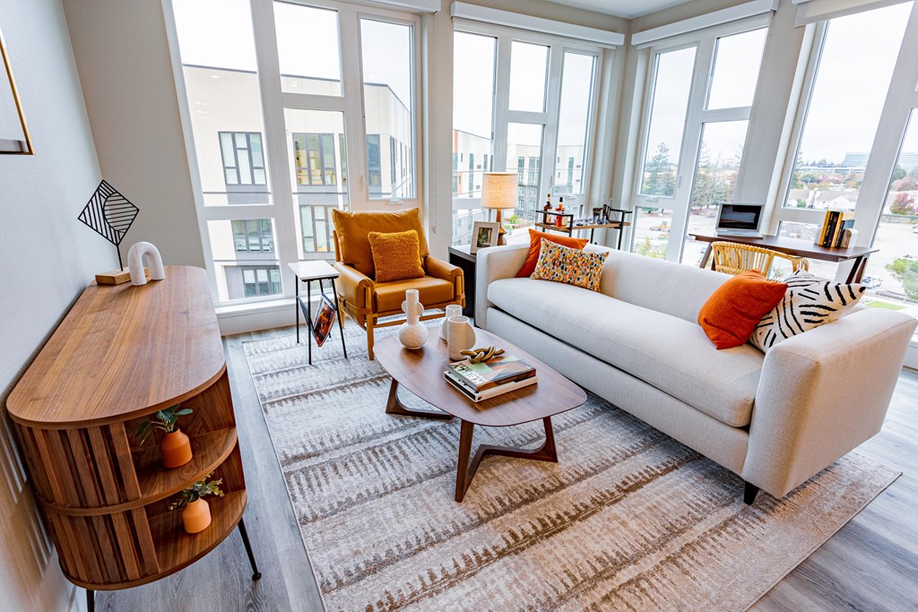Apartments in Sunnyvale for Rent - 1250 Lakeside - Living Room with Plank Style Flooring, Area Rug, Couch, Armchair, Coffee Table, Wooden Media Console, Mounted TV, Floating Shelves with Decors, and Large Window
