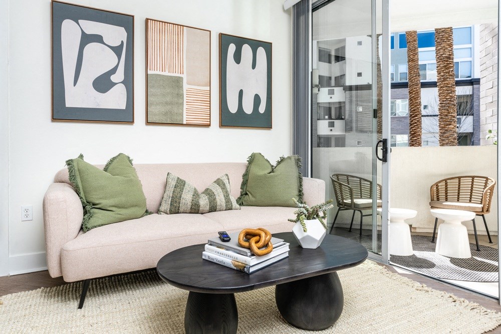 Pet Friendly Apartments in Phoenix AZ - Level at Sixteenth - Living Room with White Sofa and Green Pillows, Wood-Style Flooring, Light Area Rug, Framed Art, Dark Painted Wood Cofee Table, Balcony Access with Glass Sliding Door, and Outdoor Seating