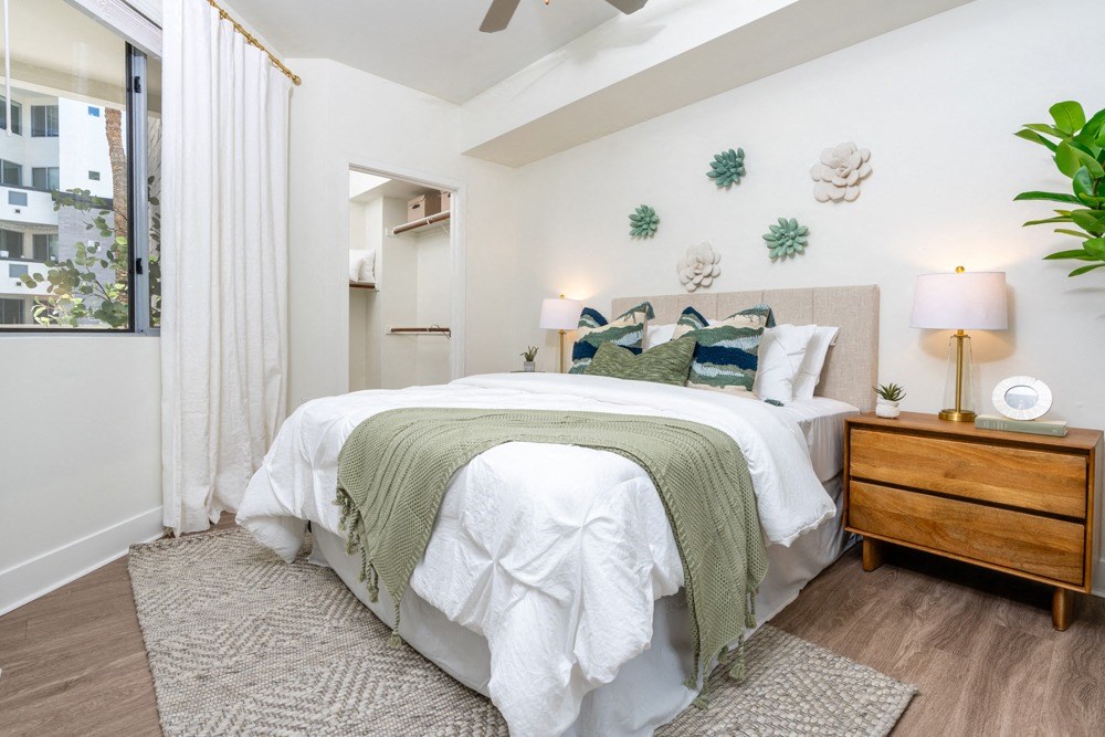One Bedroom Apartments in Phoenix AZ - Level at Sixteenth - Furnished Bedroom with Wood-Style Flooring, Area Rug, Bed, Wooden Nightstands with Lamps, White Walls, House Plant, Plant-Inspired Wall Decors, Large Closet with Hanger Bars and Storage Shelves, Window with White Curtains, and Ceiling Fan