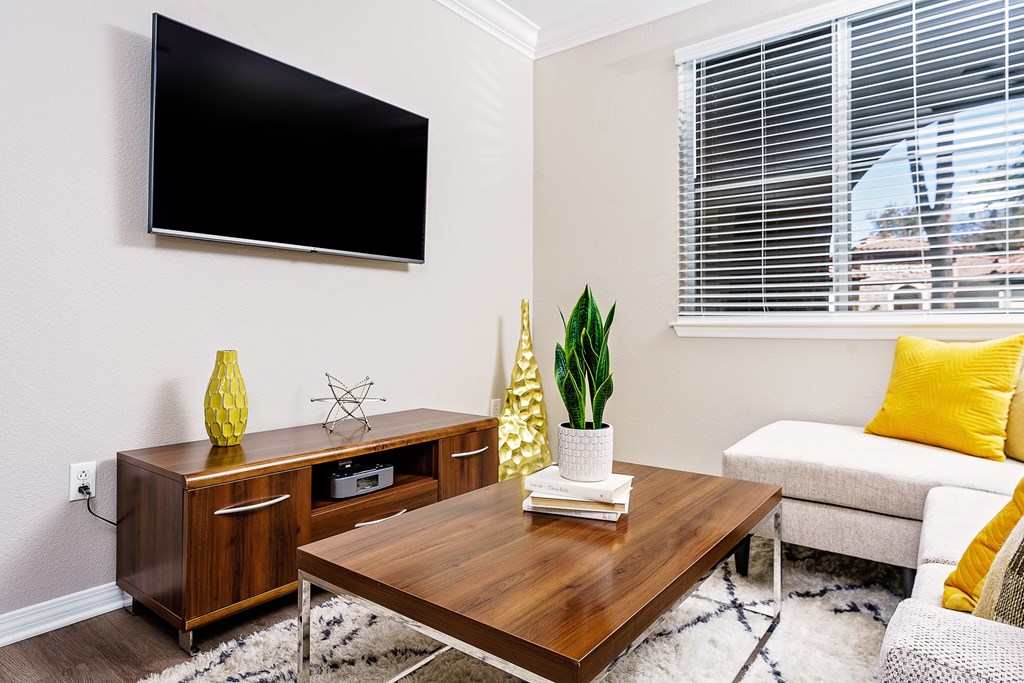 One, Two, And Three-Bedroom Apartments In Chino Hills, CA - Missions At Chino Hills - Living Room With Sectional, Coffee Table, TV Stand, TV, Plants, And Window.