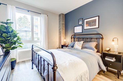 Downtown Oakland, CA Luxury Apartments Near Lake Merritt - Edson House - Bedroom With Floor Plant, Luxury Wood Finish Plank Flooring, Large Window, Concrete Pillar, Two Nightstands, and a Bed.