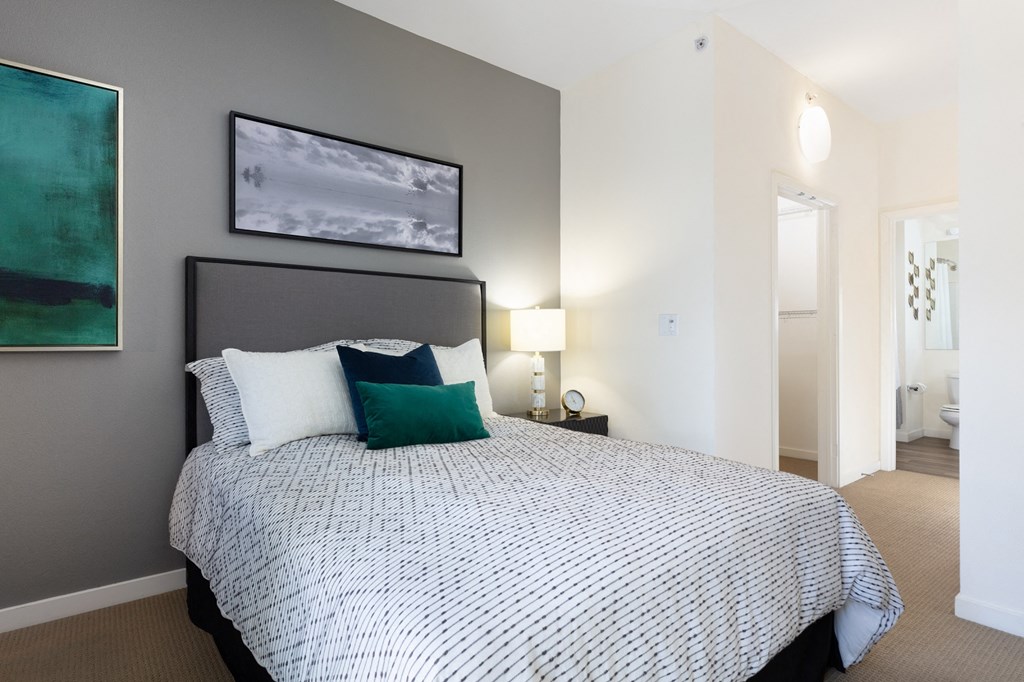 One Bedroom Apartments in Mission Bay, San Francisco CA - STRATA at Mission Bay - Furnished Bedroom with Carpeted Flooring, Bed, Nightstand with Lamp, Framed Art, Grey Accent Wall, Attached Bathroom, and Large Closet