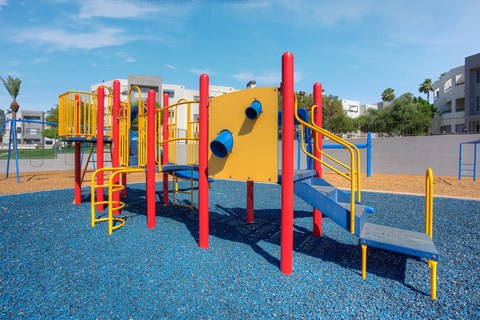 a playground with a yellow and red structure in a park at Vaseo Apartments, Phoenix, AZ High-End Apartments in North Phoenix, AZ - Vaseo - Brightly Colored Playground Structure With Jungle Gym, A Swing Set, And Monkey Bars.