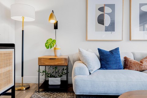 A living room with a grey couch, a wooden side table with a plant, and two framed artworks on the wall.
