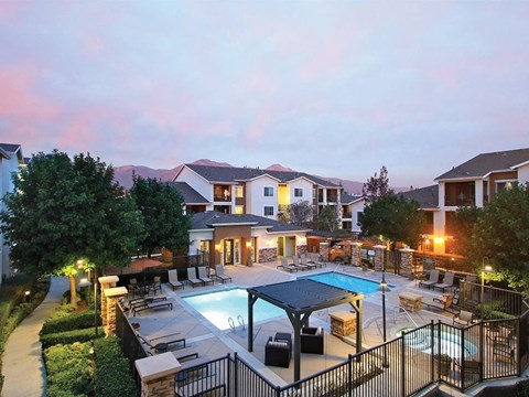 Pet-Friendly Apartments in Upland, CA - Park Central Apartments - Swimming Pool with a Spa, Sun Loungers, Lounge Seats, Pergola, and Umbrellas, Surrounded by a Metal Fence, Trees, and Landscaping.