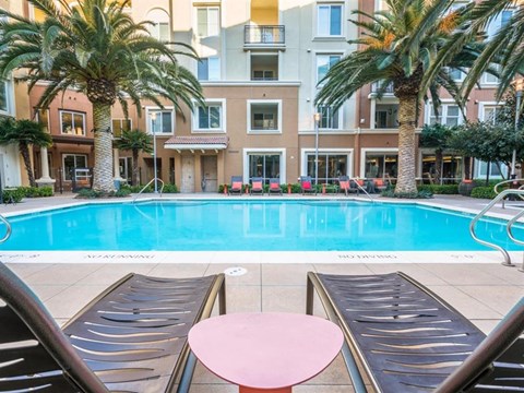 One-Bedroom Apartments in Milpitas, CA - Ilara Apartments - Swimming Pool With Lounge Chairs, Palm Trees, Clear Water, Apartment Building, Balcony Railings, And Seating Along The Deck.