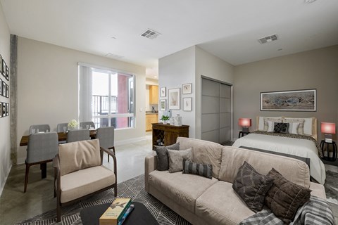 2-Bedroom Modern Apartments in Long Beach, CA - Urban Village - Open Living Room With Sofa Seating, Dining Area, Bed, Large Window, And Bright Neutral Interior.