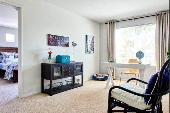Tustin, CA, Townhomes Near Santa Ana - Tustin Cottages - Living Room With Desk, Console Cabinet, Armchair, Pet Bed, Large Window, Neutral Carpet, and Adjacent Bedroom Entry.