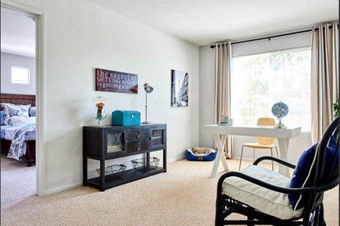 Tustin, CA, Townhomes Near Santa Ana - Tustin Cottages - Living Room With Desk, Console Cabinet, Armchair, Pet Bed, Large Window, Neutral Carpet, and Adjacent Bedroom Entry.