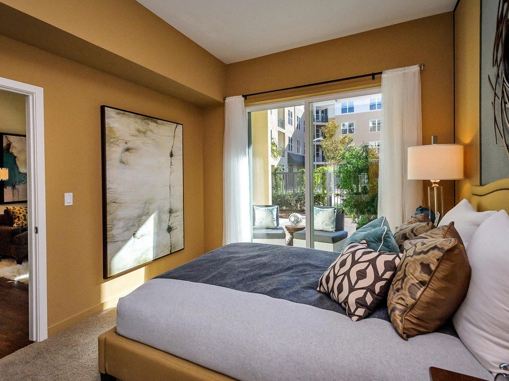 Three-Bedroom Apartments in Foster City, CA - The Plaza - Bedroom with Carpeting, Bed Standing Lamp, Wall Art, and Sliding Doors with Access to Patio