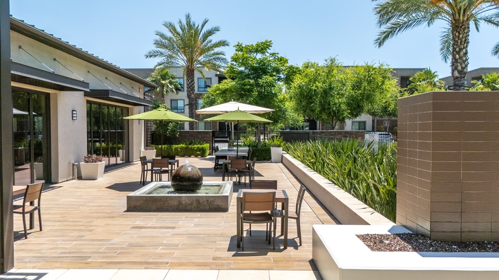 A patio with a fountain and chairs is surrounded by palm trees.