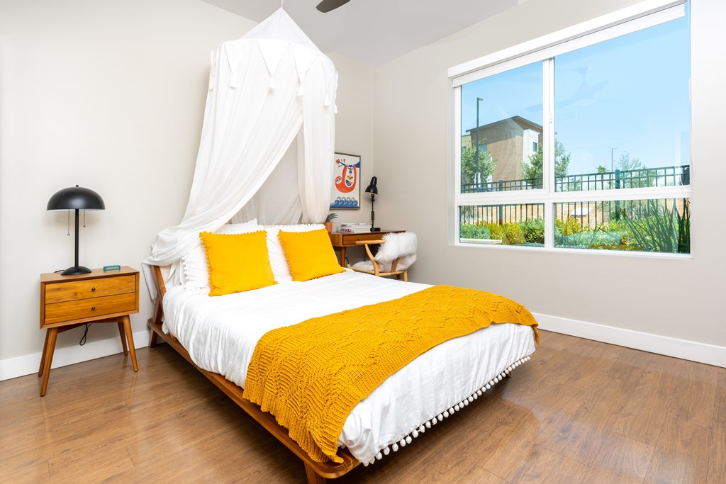 A bedroom with a canopy bed, a nightstand, and a window overlooking a fence.