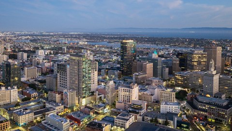 A cityscape at dusk with buildings illuminated and a body of water in the distance.