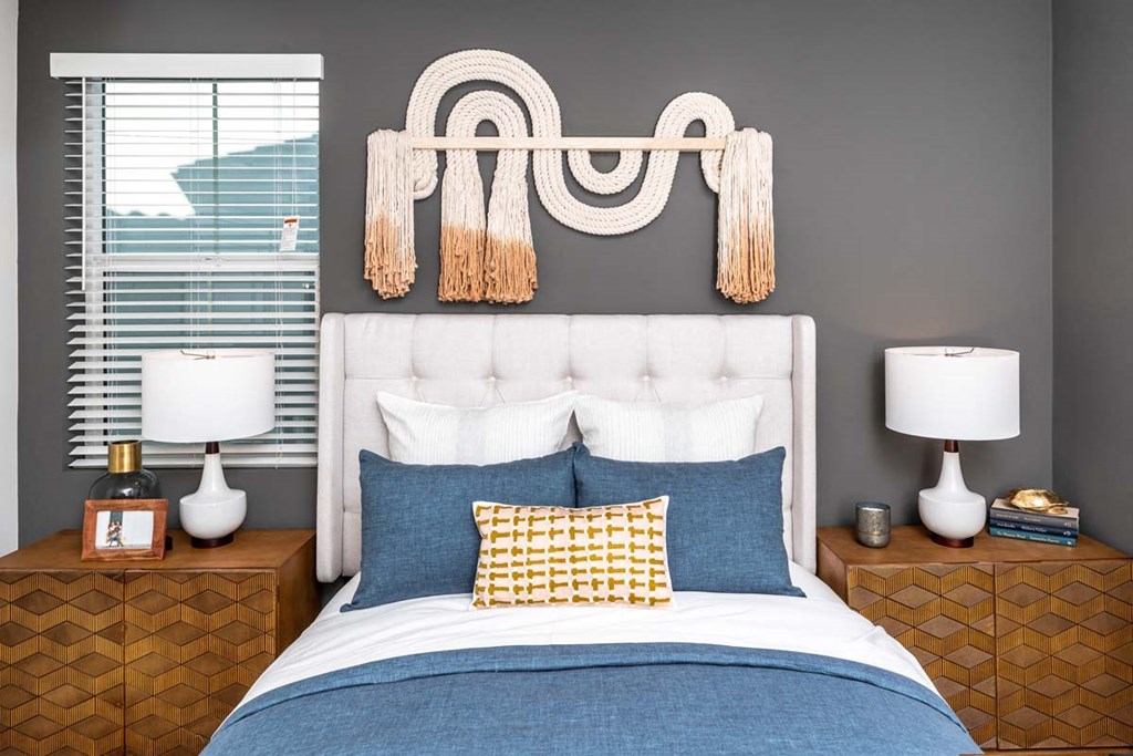 Luxury One-Bedroom Apartments in Corona, CA - The Monterey Corona - Furnished Bedroom with Grey Walls, Carpeted Flooring, Bed, Wooden Nightstands with Lamp, Wicker Chair, Hanging Wall Art, and Two Windows with Blinds and White Curtains