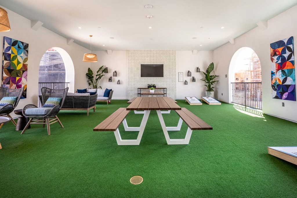 Apartments in Corona CA - The Monterey Corona - Recreational Area with Artificial Turf, Picnic Benches, Cushioned Chairs, Sofas, Plants, Framed Wall Art, Corn Hole Boards, TV, and Overhead Lights