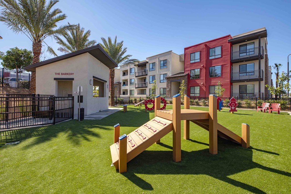 Pet-Friendly Apartments in Redlands, CA - The Summit - Dog Park With Grass Turf, Pet Waste Station, Agility Equipment, Lush Landscaping, and Red Garden Patio Chairs