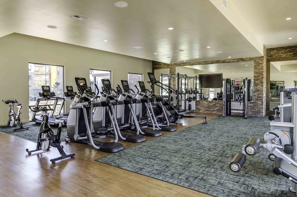 Apartments for Rent in Redlands, CA - The Summit - Fitness Center With Large Windows, Cardio Equipment, Weight Training Machines, Puck Lighting, Mirrors, Sectioned Off Carpeting, Mounted TV, and Wood-Style Flooring